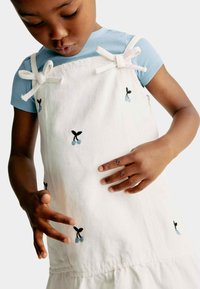 Hvid denim samlede kjole med blå kirsebærbroderi, justerbare hvide sløjfesele, lagdelt flæsekanter, båret over en lys blå ribbet skjorte.