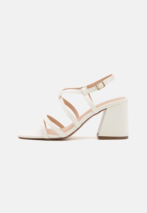 Sandale blanche à brides avec talon bloc, boucle de cheville ajustable, et bout ouvert sur un fond clair uni.