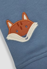 Broderet orange og hvid rævejævn med syede øjne og næse på blåt stof, der viser teksturerede strikkede detaljer.