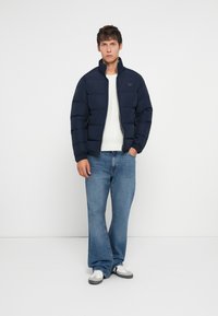 Young man wearing a navy puffer jacket, white sweater, blue jeans, and white sneakers, standing with one hand in pocket against plain background.