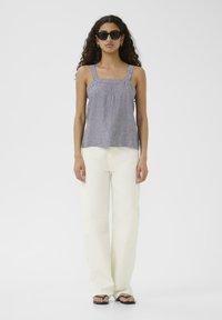 Bez rękawów, jasnoszara bluzka z kwadratowymi ramiączkami i marszczonym przodem; w połączeniu z ecru spodniami o szerokich nogawkach i czarnymi sandałami.