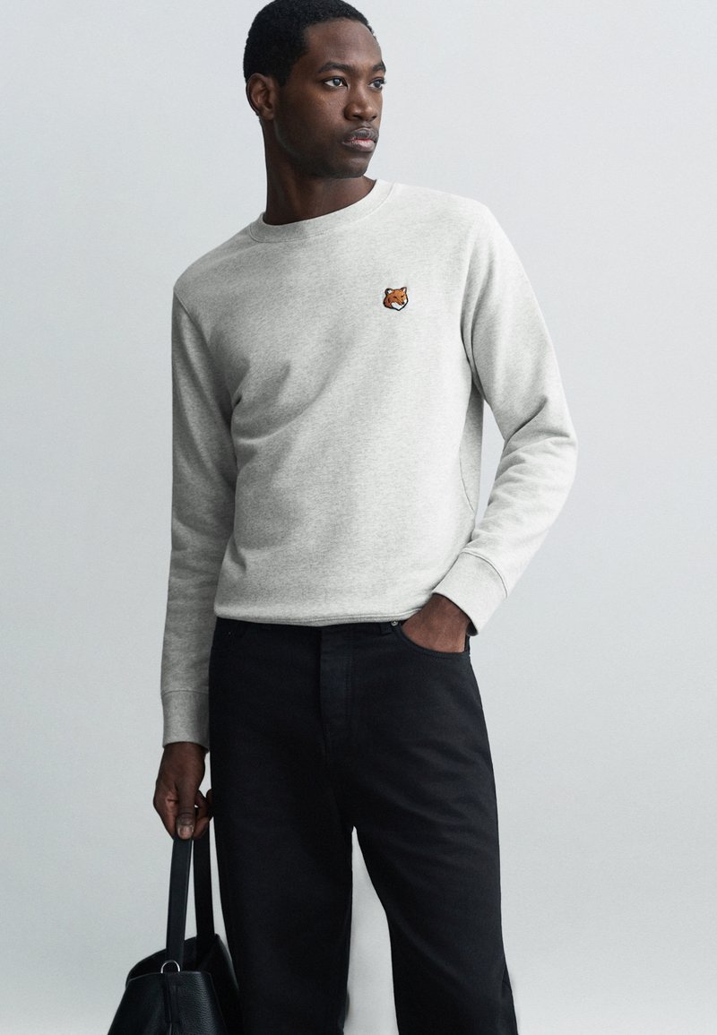 Sweatshirt in felpa grigia con maniche lunghe, scollo rotondo e piccolo design di un animale ricamato; abbinato a pantaloni neri e una borsa nera.