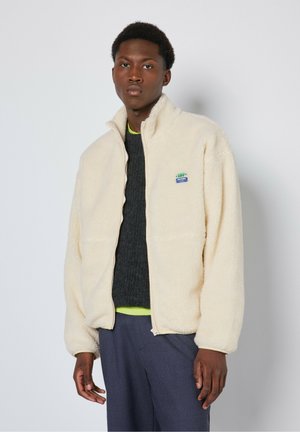 Veste en polaire couleur crème avec un col haut, fermeture éclair, deux poches avant et une petite étiquette logo bleue sur la poitrine.