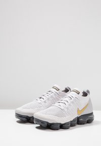Witte Nike Air Max-schoenen met een mesh bovenwerk, gouden accenten en een chunky, transparante zool. Heeft een gestructureerd patroon op het gehele oppervlak.