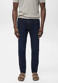 Jeans de denim azul oscuro con corte ajustado, que cuentan con bolsillos estándar y detalles de costura clara, combinados con sandalias marrones.
