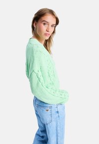 Minzgrüner, kabelgestrickter Pullover mit lockerer Passform und überschnittenen Schultern, kombiniert mit hellblauen High-Waist-Jeans. Strukturierter Stoffdetails sichtbar.