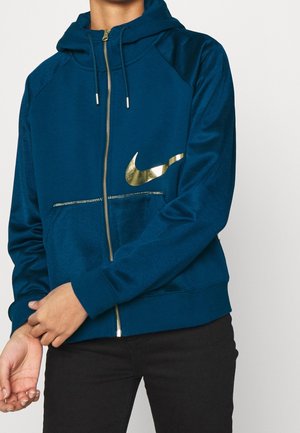 Personne portant un sweat à capuche bleu zippé avec un logo Nike doré et un pantalon noir, ajustant la zone du poignet de la manche.