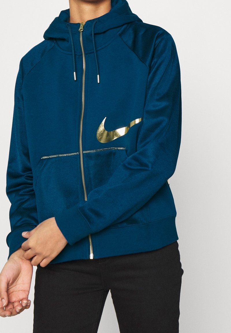 Personne portant un sweat à capuche bleu zippé avec un logo Nike doré et un pantalon noir, ajustant la zone du poignet de la manche.