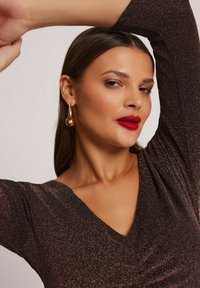 Błyszcząca, ciemnoczekoladowa sukienka z dekoltem w serek i rękawami do trzech czwartych. Modelka ma na sobie złote kolczyki w kształcie kropli i ciemnoczerwony błyszczyk do ust. Gładka tekstura.