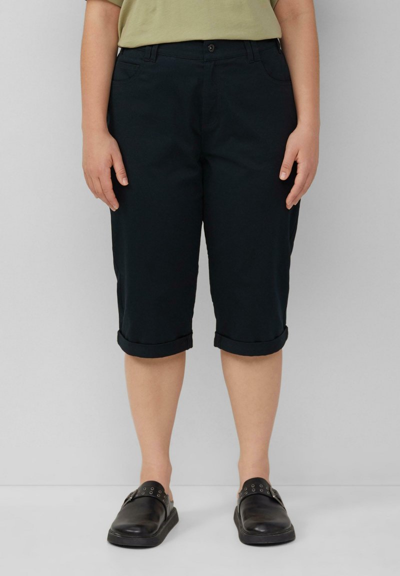 Pantaloni neri al ginocchio con risvolti, realizzati in tessuto morbido. Presenti tasche frontali e passanti per cintura. Indossati con scarpe nere senza lacci.