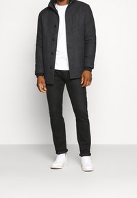 Uomo che indossa un cappotto grigio scuro con bottoni, una maglietta bianca, pantaloni neri e sneakers bianche, in piedi contro uno sfondo chiaro e semplice.
