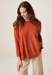 Rostfarbener Strickpullover mit weitem Ärmel und geripptem Saum. Glatte Textur, lockere Passform, kombiniert mit hellen Hosen.