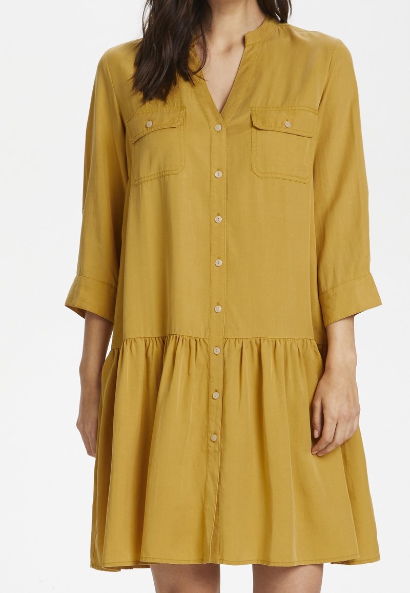 Robe jaune moutarde avec un devant boutonné, deux poches poitrine et un ourlet évasé. Elle présente des manches trois-quarts et un tissu doux et léger.