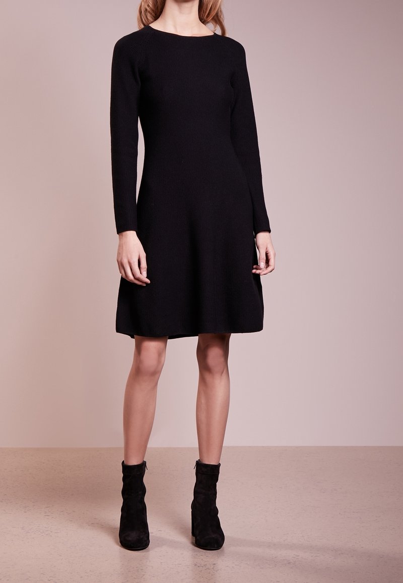 Femme portant une robe noire en maille à manches longues associée à des bottines noires, debout devant un fond beige uni.