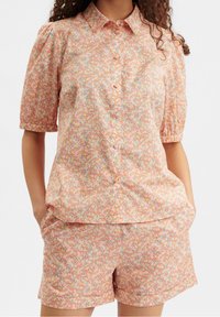 Set di camicia e pantaloni corti a fantasia floreale in pesco, con maniche corte, chiusura a bottoni e tasche. Il tessuto sembra leggero e testurizzato.