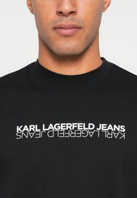 Melna t-krekls, kas izgatavots no gludas auduma, ar izteiksmīgu baltu tekstu, kas horizontāli drukāts pāri krūtīm un lasāms "KARL LAGERFELD JEANS".