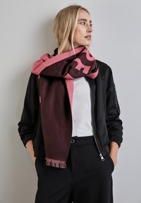 Maronfarvet og rosa tørklæde med ternet mønster og frynsede kanter, båret over en sort jakke, der har en hyggelig tekstur og lagdelt fald.
