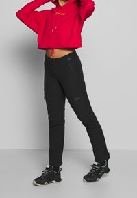 Sudadera corta roja con cordones, pantalones ajustados negros con bolsillos con cremallera, y zapatillas de senderismo negras con detalles en azul verdoso.