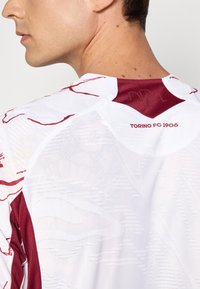 Fehér sportmez bordó részletekkel, diszkrét mintákkal, sima textúrával, és a nyak mögött a "TORINO FC 1906" felirattal.