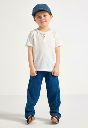 Ung dreng står, iført en blå kasket, hvid kortærmet skjorte, blå jeans og sorte sandaler på en ensartet lys baggrund.