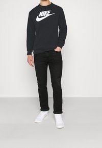 Musta huppari, jossa on valkoinen Nike-logo, yhdistettynä mustiin farkkuihin ja valkoisiin lenkkareihin. Siinä on pyöreä pääntie ja ribbaatut hihansuut.