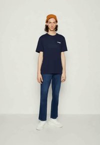 T-shirt de algodão azul navy com mangas curtas e logótipo no peito, combinada com jeans de ganga azuis e ténis brancos. Modelo a usar um gorro laranja.
