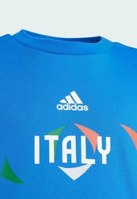T-shirt in cotone blu con la scritta bianca "ITALY", logo Adidas e accenti geometrici colorati in verde, arancione e bianco. Design con colletto a girocollo.