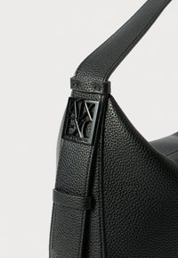 Borsa in pelle nera con texture, caratterizzata da una forma curvata, una tracolla regolabile e un'etichetta con logo nero ben visibile.