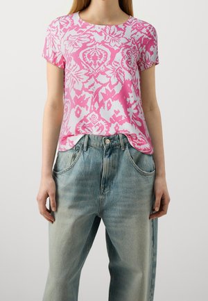 Persoon draagt een roze-wit shirt met bloemenpatroon en korte mouwen, ingestopt in losse lichtblauwe jeans, met handen langs de zij.