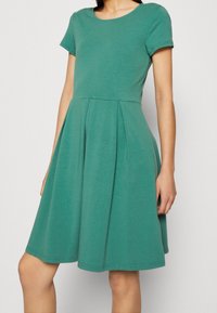 Kortärmad klänning i teal bomull med rund halsringning, figurnära bodice och utsvängd kjol. Har vertikala veck vid midjan för extra detalj.