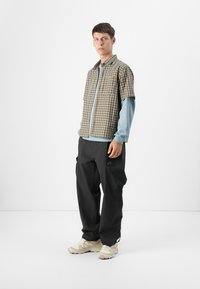 Young male model stands in a minimalist setting, wearing a checkered short-sleeve shirt over a long-sleeve blue top and black pants.