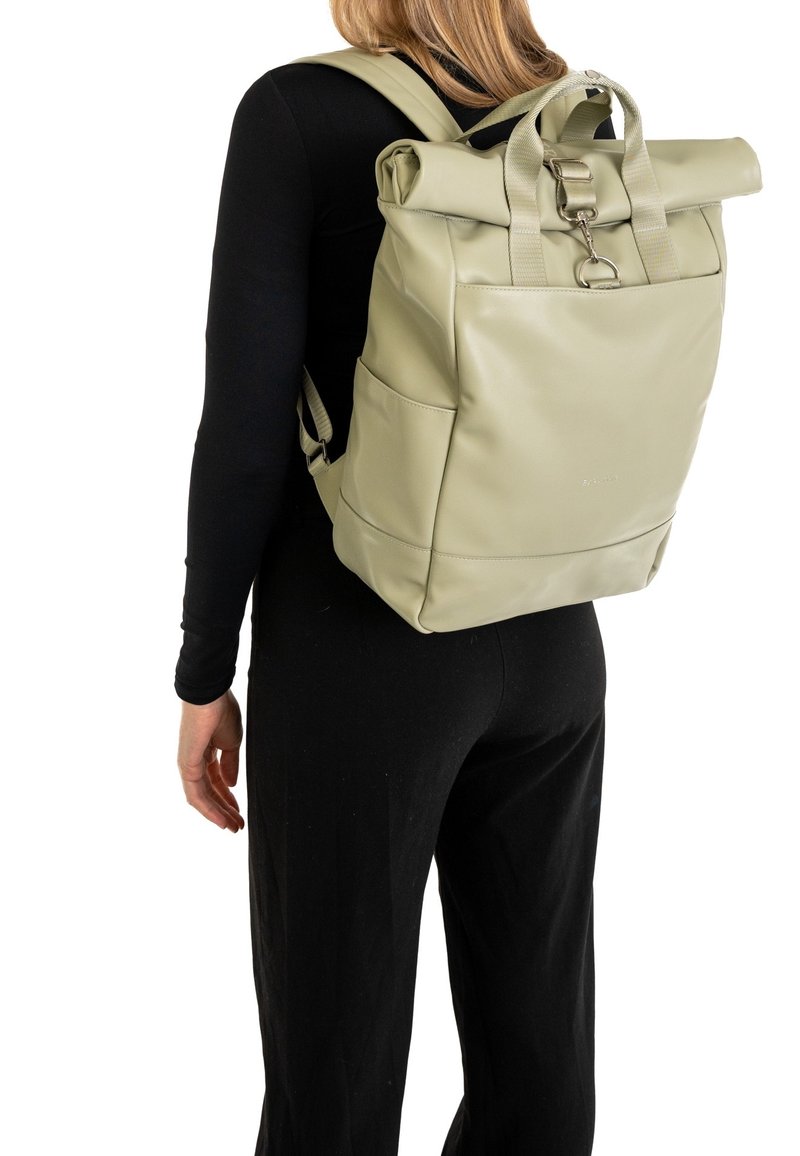Personne portant un sac à dos roll-top vert clair avec poche avant et poches latérales, habillée de manches longues noires et pantalon, debout face à nous mais de dos.