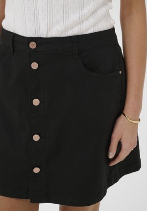 Femme portant une jupe noire à boutons sur le devant avec des poches, associée à un haut blanc à motifs et un bracelet doré au poignet droit.