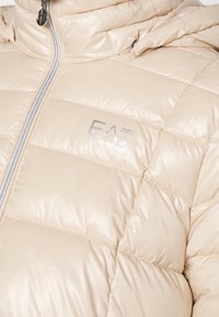 Beżowa kurtka puchowa z srebrnym zamkiem i logo EA7 Emporio Armani na klatce piersiowej, z pikowanym ściegiem i wysokim kołnierzem.