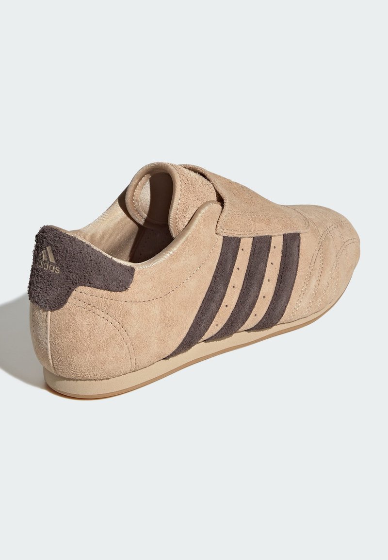 Zapatilla deportiva de ante en beige con acentos de rayas marrón oscuro. Presenta una punta redondeada, un cuello acolchado y un parche texturizado en el talón.
