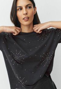 Zwart t-shirt met korte mouwen van katoen, voorzien van verspreide metalen versieringen die een stervormig patroon creëren. Soepele textuur en een ontspannen pasvorm.