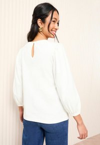 Blouse blanche avec des manches bouffantes, encolure ronde et détail en forme de clé à l'arrière, associée à un jean en denim bleu. Texture lisse, design simple.