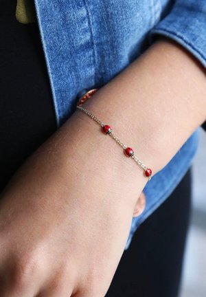Gouden kettingarmband met drie rode kralen met zwarte accenten, gedragen om een pols. Achtergrond bestaat uit denimstof en huidskleur.