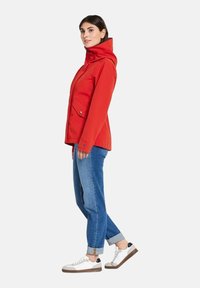 Veste imperméable rouge avec un col montant, des poches avant et une fermeture éclair, assortie à un jean bleu clair retroussé et des baskets blanches.