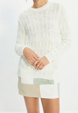 Femme portant un pull en maille torsadée blanc et une mini-jupe patchwork en velours côtelé beige, blanc et vert clair, les mains jointes devant.