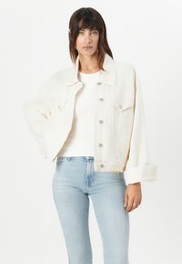 Veste en denim blanche avec un col, des boutons à l'avant et deux poches poitrine. Portée par-dessus un haut blanc à côtes et un jean bleu clair.