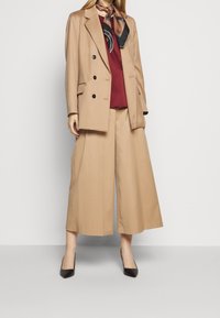 Blazer doppiopetto beige, top bordeaux e pantaloni beige a gamba larga. Tacchi neri a punta e una sciarpa beige annodata completano l'outfit.