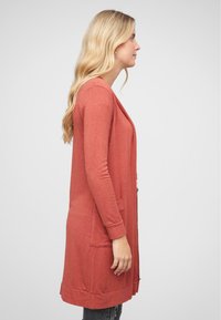 Lang cardigan i rustfarge, laget av mykt strikkemateriale. Har åpen front, to sidelommer og ribbestrikkede mansjetter og kant.