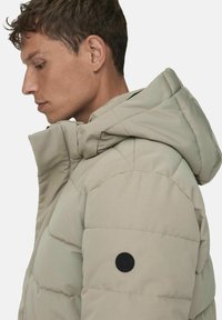 Vaalean beige-puffatakki hupulla, tikattu pinta ja pyöreä musta logo hihassa. Sivuprofiilista näkyvät muotoilu ja yksityiskohdat.