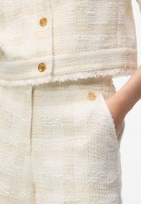 Tenue en tissu texturé blanc avec un motif à carreaux. La veste courte est ornée de boutons dorés. Le pantalon présente des détails assortis et des poches.