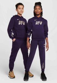 Sudaderas con capucha moradas y pantalones con el logo blanco "KM". Los pantalones tienen un diseño de llamas en los puños. El calzado del modelo incluye zapatillas negras y doradas.