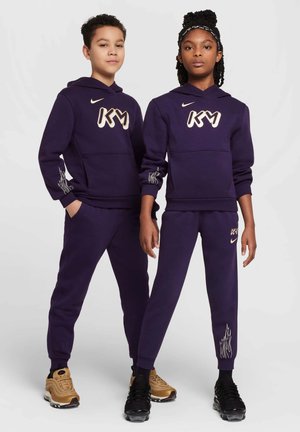 Felpe con cappuccio viola e pantaloni con logo bianco "KM". I pantaloni presentano un design a fiamma sui polsini. Le scarpe del modello includono sneakers nere e dorate.
