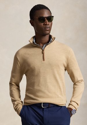 Homme portant des lunettes de soleil, un pull beige à fermeture éclair jusqu'au quart sur une chemise rayée, un pantalon bleu marine et une montre-bracelet, les mains dans les poches.