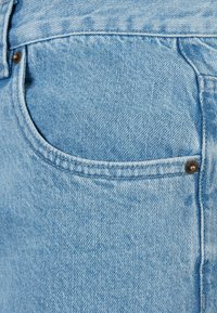 Gros plan sur la poche d'un jean en denim bleu clair avec des surpiqûres visibles et des rivets métalliques sur la texture du tissu.