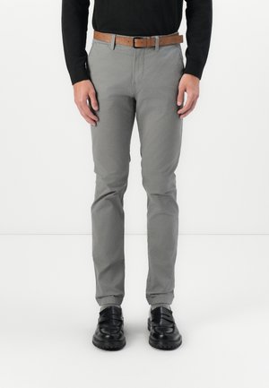 Mann trägt graue Slim-Fit-Hose mit braunem Gürtel, schwarzen Loafers und schwarzem Langarmshirt, steht vor weißem Hintergrund.